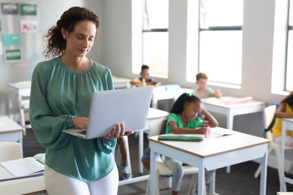 Junge Lehrerin steht im Klassenzimmer und hält einen Laptop in der Hand, während die Kinder im Hintergrund an den Tischen sitzen und demonstrieren, wie Lehrer KI-Tools im Unterricht einsetzen