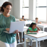 若い女性教師が教室でノートパソコンを持ち、机に向かっている子どもたちを背景に立っている。