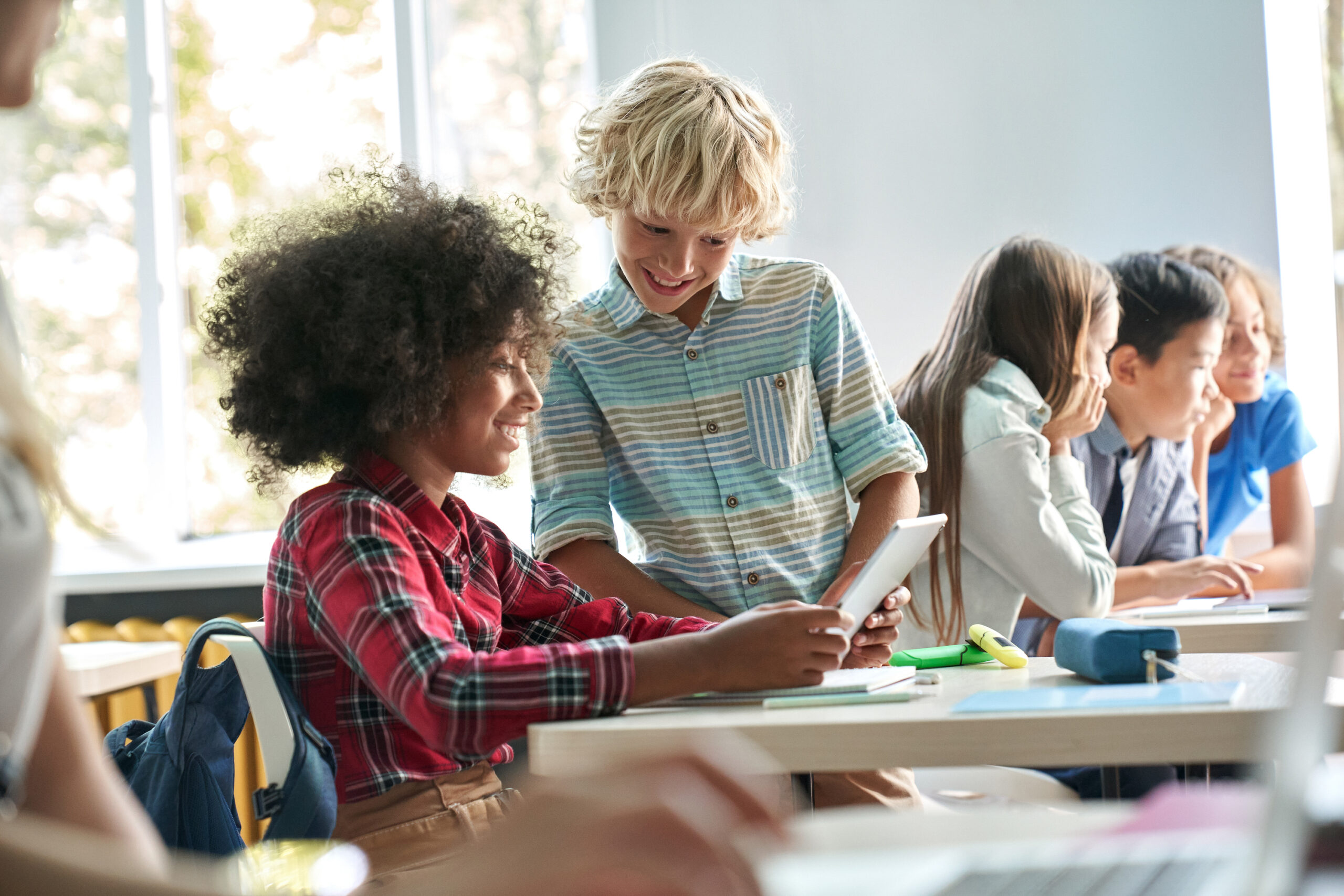 Happy smiling Caucasian boy and African American girl schoolchildren studying together using tablet device in classroom. Groups of schoolchildren working on task. Using technologies for education showing the concept of digital education access