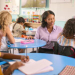 小学校または小学校の女性教師が、多文化クラスで机に向かっている生徒を助け、評価ツールの使い方を示している。