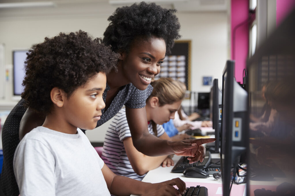 Female teacher leaning over a young male student sitting in a computer class that has implemented digital assessment strategies