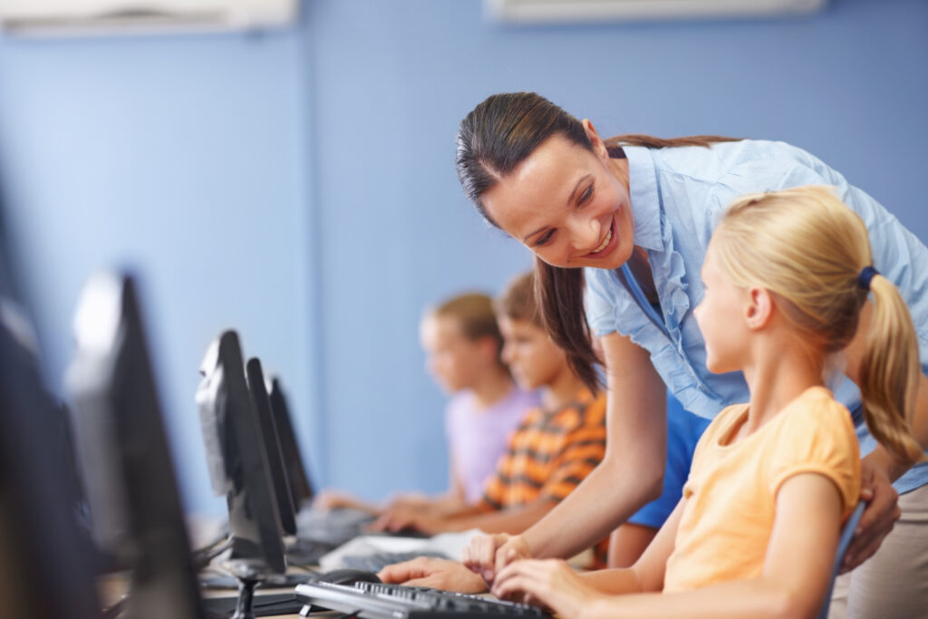 Young female teacher leaning over a young female student sitting at a computer and using a learning assessment tool