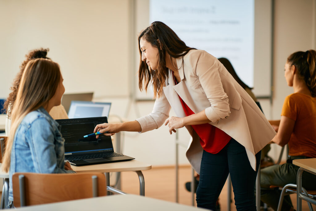 Young female teacher in a classroom leaning over a female student's desk and pointing at her computer demonstrating the concept of benchmark assessments