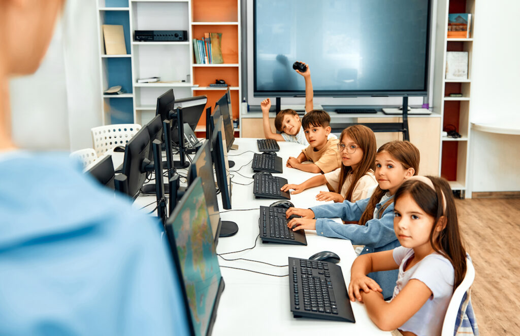 School children sitting in a row in computer class facing their computers and looking off to the side at teacher, illustrating the concept of comprehensive assessment.