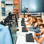 School children sitting in a row in computer class facing their computers and looking off to the side at teacher, illustrating the concept of comprehensive assessment.