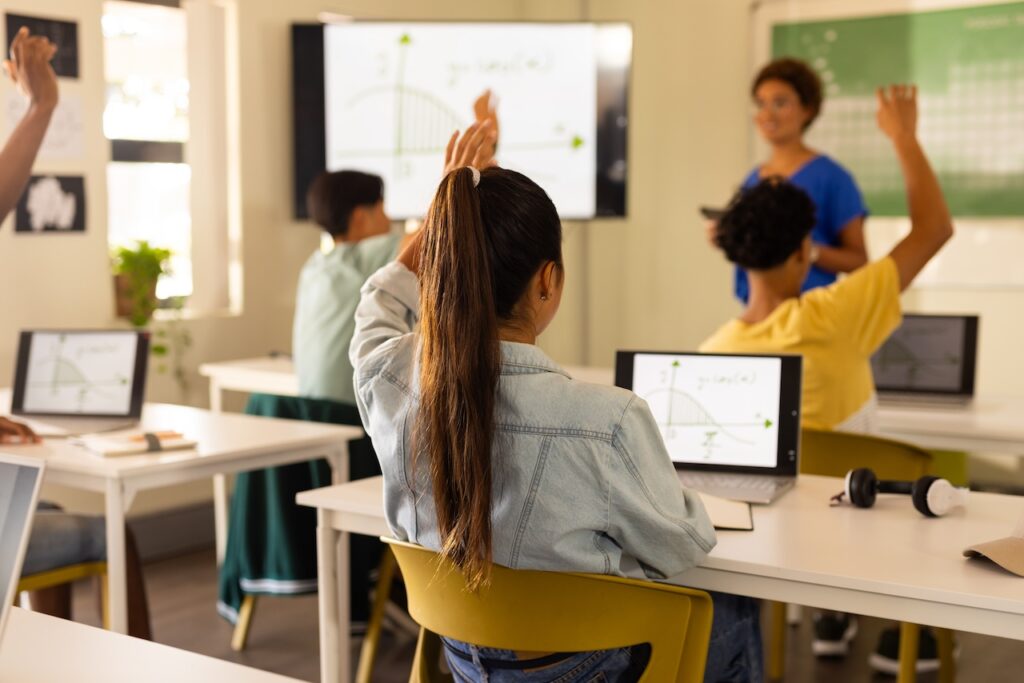 View from behind of girl sitting in a classroom at a desk in front of a laptop with a math equation on the screen, raising her hand as the teacher presents at the front of the class, demonstrating the concept of diagnostic assessments in math.