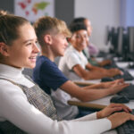 Close up side view of male and female students sitting in a row at computers, smiling at the screen with their hands on the keyboard, demonstrating an example of a formative assessment being taken.