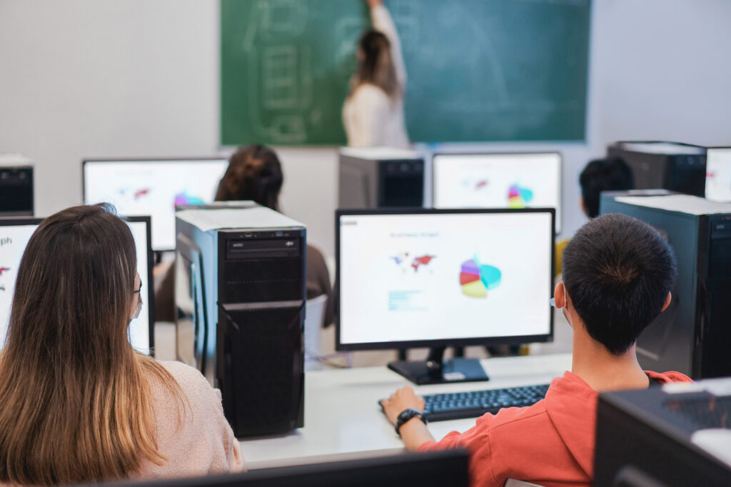 Rear view of students in class sitting at desks in front of computers facing a female teacher with her back turned drawing on a chalkboard, illustrating students taking formative or summative assessments.