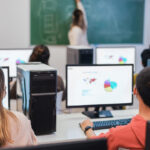 Rear view of students in class sitting at desks in front of computers facing a female teacher with her back turned drawing on a chalkboard, illustrating students taking formative or summative assessments.