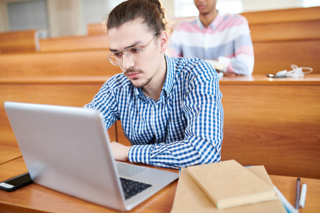 College age male student sitting in a lecture hall taking a summative evaluation on his laptop.