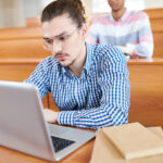 College age male student sitting in a lecture hall taking a summative evaluation on his laptop.