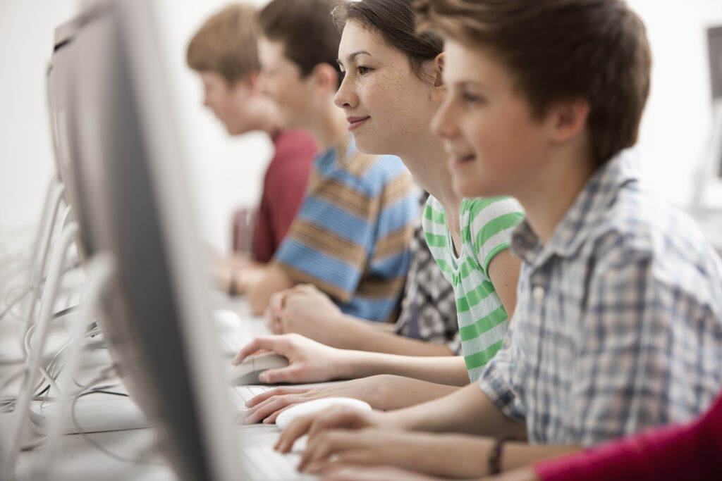 Profile view of four children sitting in a row of desks typing on school computers while taking adaptive tests.