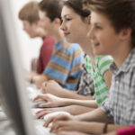 Profile view of four children sitting in a row of desks typing on school computers while taking adaptive tests.