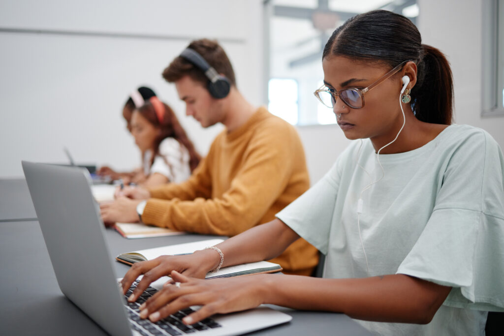 Side view of three students sitting in a row in front of their computers and wearing headphones taking an online assessment