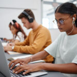 Side view of three students sitting in a row in front of their computers and wearing headphones taking an online assessment