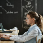 Side view of school girl sitting at a desk in front of a chalkboard facing her laptop.